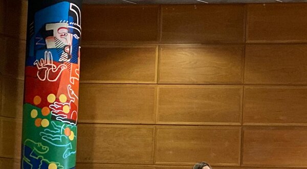 Mulher a falar num pódio, com painel colorido ao fundo e público sentado.