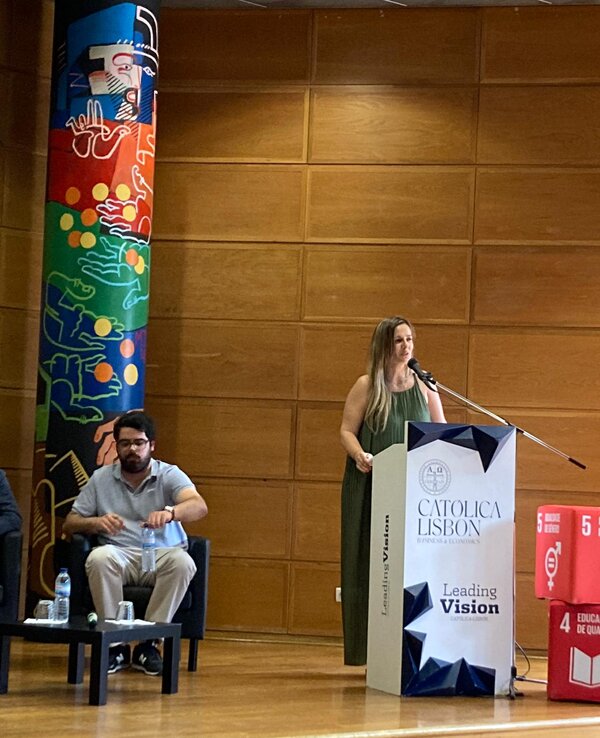 Mulher a falar num pódio, com painel colorido ao fundo e público sentado.