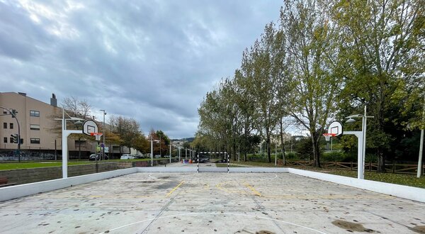 Campo de basquetebol ao ar livre, com cestos e árvores ao fundo.