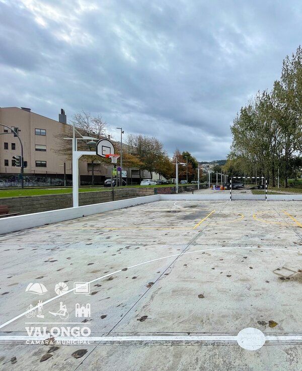 Campo de basquetebol ao ar livre, com cestos e árvores ao fundo.