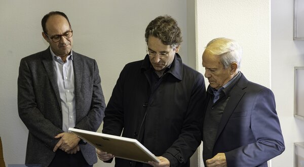 Três homens em um ambiente interno, observando um quadro.