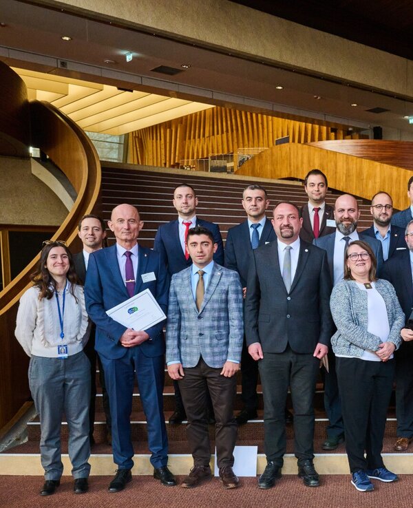 Grupo de pessoas em cerimónia, segurando certificados, em escadaria elegante.