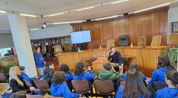 Grupo de crianças em uma sala, ouvindo um orador em um painel.