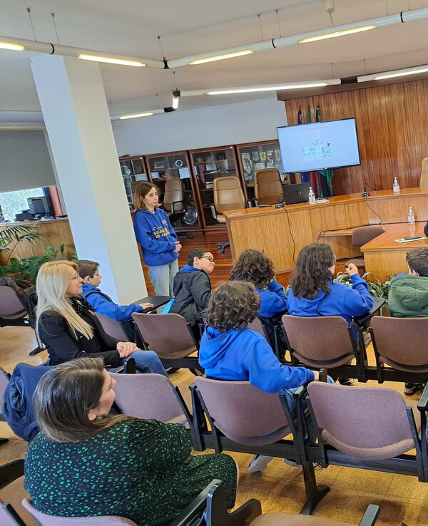 Grupo de crianças em uma sala, ouvindo um orador em um painel.