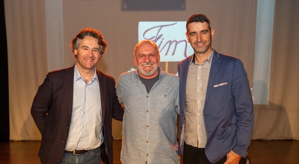 Três homens posam juntos em um palco, com um fundo neutro.