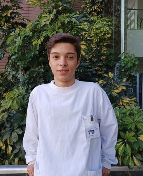 Jovem em bata branca, com crachá, em frente a plantas verdes.