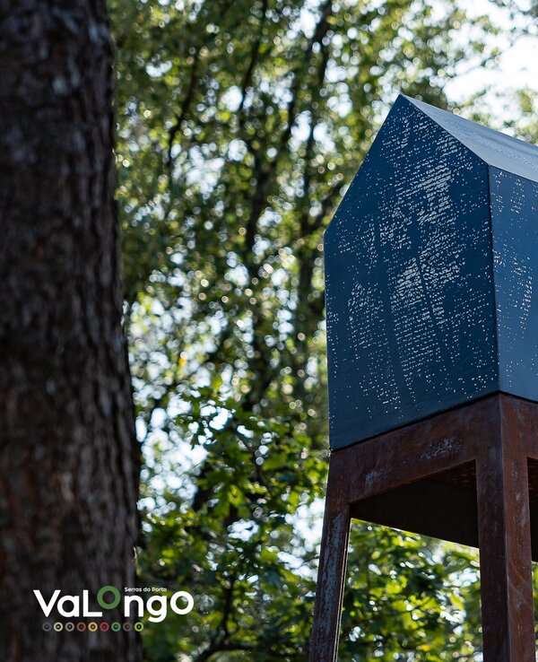 Estrutura metálica em forma de casa, elevada sobre um suporte, rodeada por árvores.
