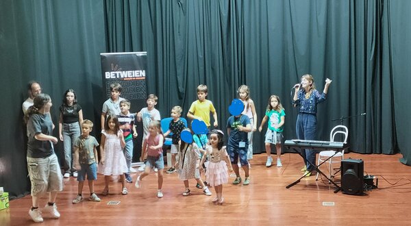 Grupo de crianças a atuar num palco, com uma mulher a cantar ao microfone.