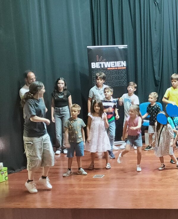 Grupo de crianças a atuar num palco, com uma mulher a cantar ao microfone.