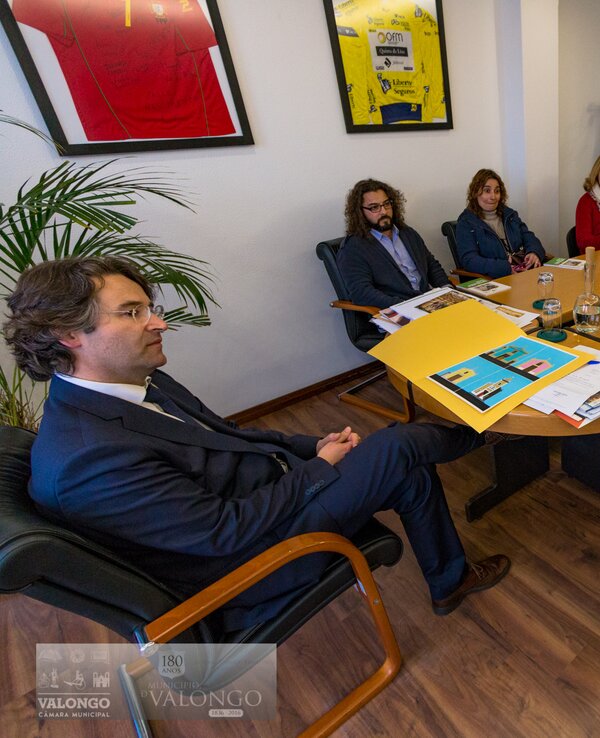 Reunião em mesa oval com várias pessoas e documentos sobre a mesa.