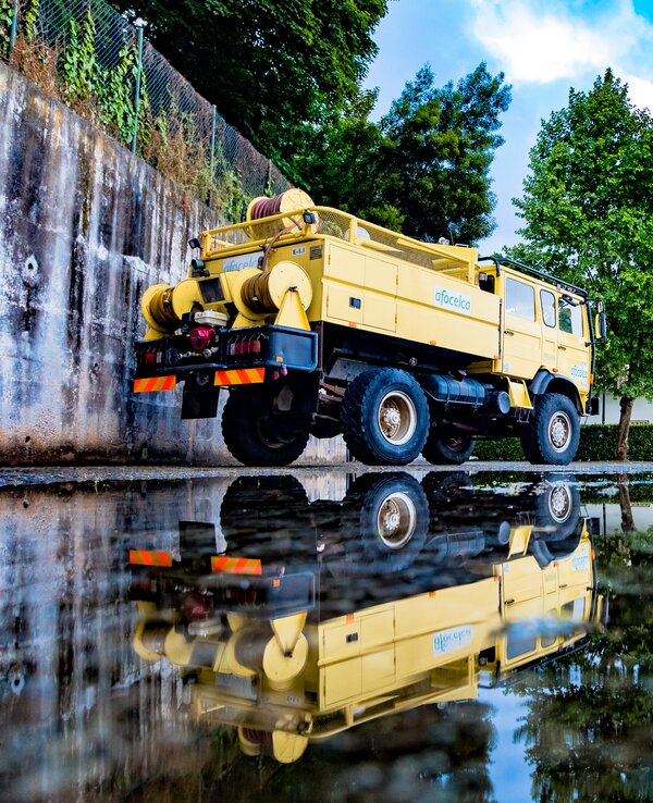 Caminhão amarelo e veículos de bombeiros refletidos em poça de água.