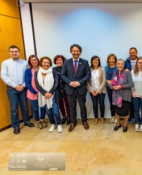 Grupo de pessoas posando em frente a um ecrã, com bandeiras ao fundo.