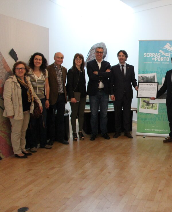 Grupo de pessoas posando em sala com painel informativo e certificado.