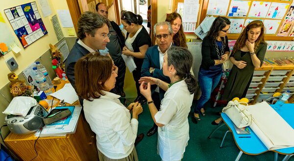 Grupo de pessoas conversando numa sala, com mesas e materiais escolares ao fundo.