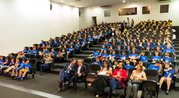 Grande auditório com crianças em camisetas azuis sentadas em filas.