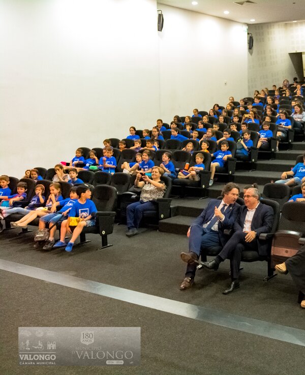 Grande auditório com crianças em camisetas azuis sentadas em filas.