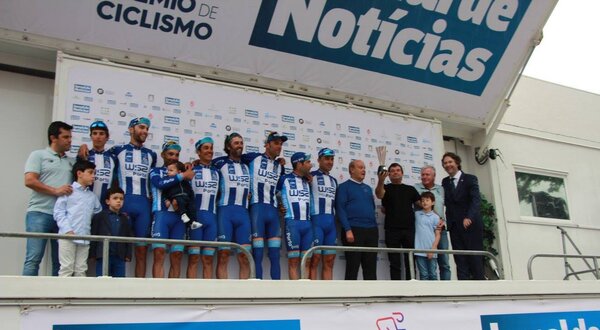 Equipa de ciclismo em palco, com patrocinadores e crianças, em evento desportivo.