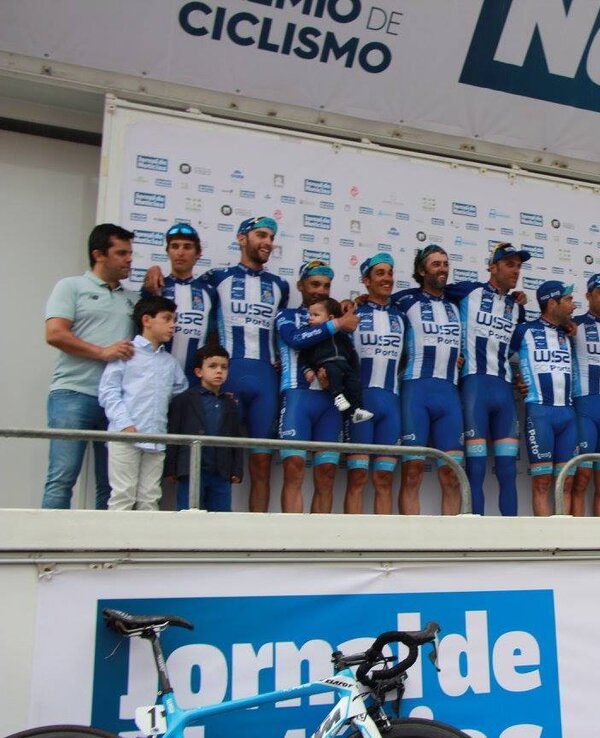 Equipa de ciclismo em palco, com patrocinadores e crianças, em evento desportivo.
