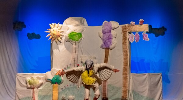 Ator em palco com cenário colorido de árvores e sol, representando uma peça de teatro.