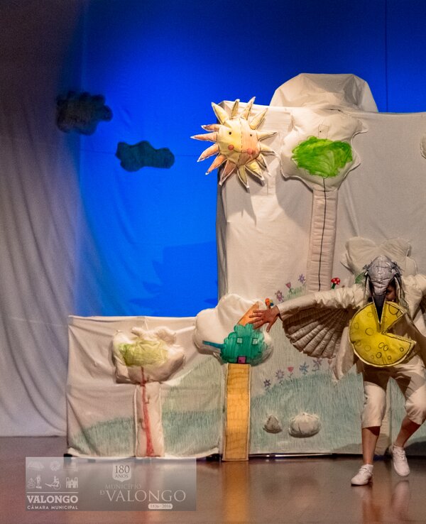 Ator em palco com cenário colorido de árvores e sol, representando uma peça de teatro.