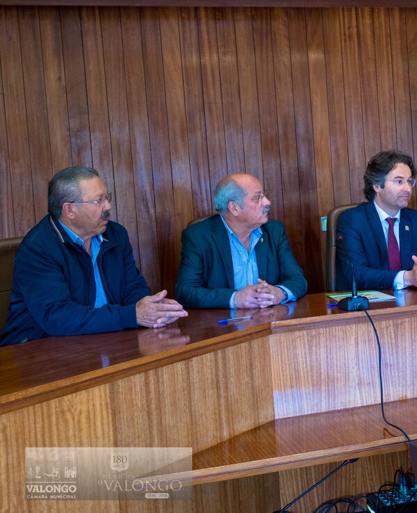 Cinco homens e uma mulher sentados à mesa em uma reunião, com bandeiras ao fundo.
