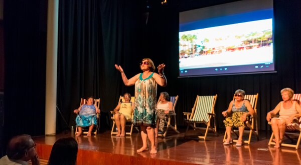 Uma mulher em destaque a atuar num palco, rodeada por outras sentadas.