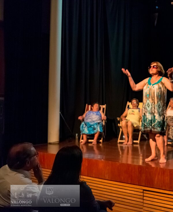 Uma mulher em destaque a atuar num palco, rodeada por outras sentadas.