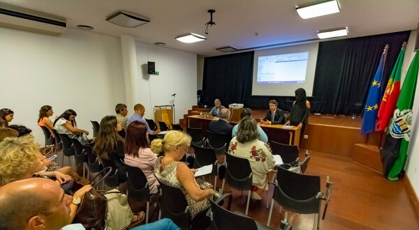Reunião com várias pessoas sentadas, apresentador em destaque ao fundo.