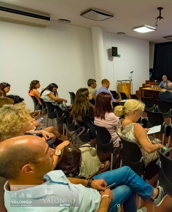 Reunião com várias pessoas sentadas, apresentador em destaque ao fundo.