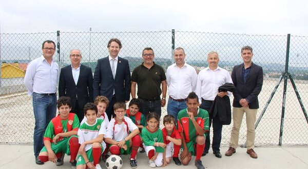 Grupo de crianças em uniforme de futebol com adultos ao fundo, em campo.