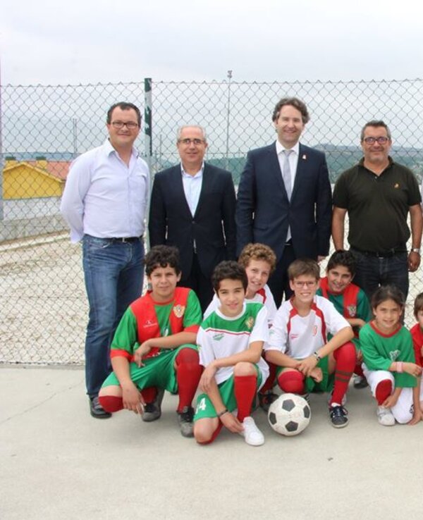 Grupo de crianças em uniforme de futebol com adultos ao fundo, em campo.
