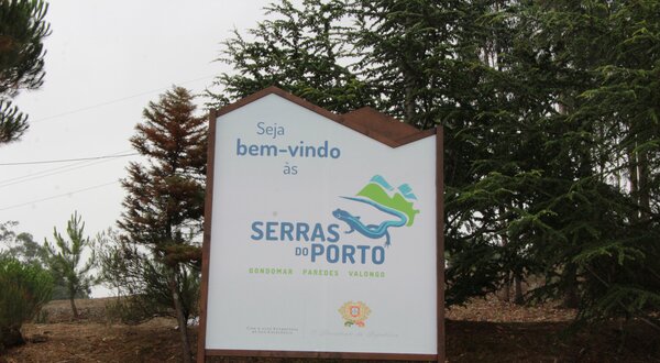 Placa de boas-vindas às Serras do Porto, com árvores ao fundo.