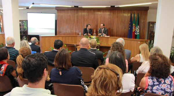 Reunião com várias pessoas sentadas, dois oradores ao fundo.