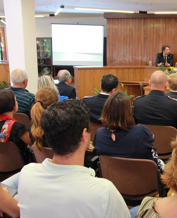 Reunião com várias pessoas sentadas, dois oradores ao fundo.
