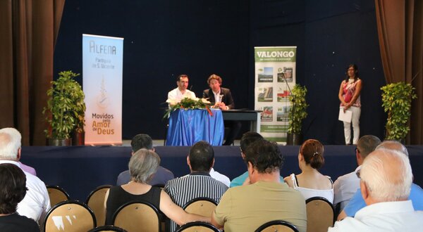 Reunião com público sentado, oradores à mesa e cartazes ao fundo.