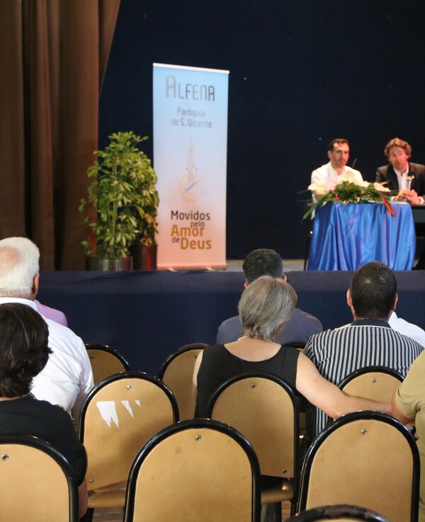 Reunião com público sentado, oradores à mesa e cartazes ao fundo.