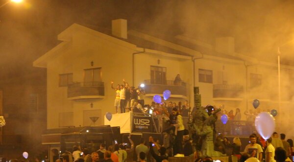 Multidão festiva à noite, com balões e fumaça, celebrando em frente a um edifício.