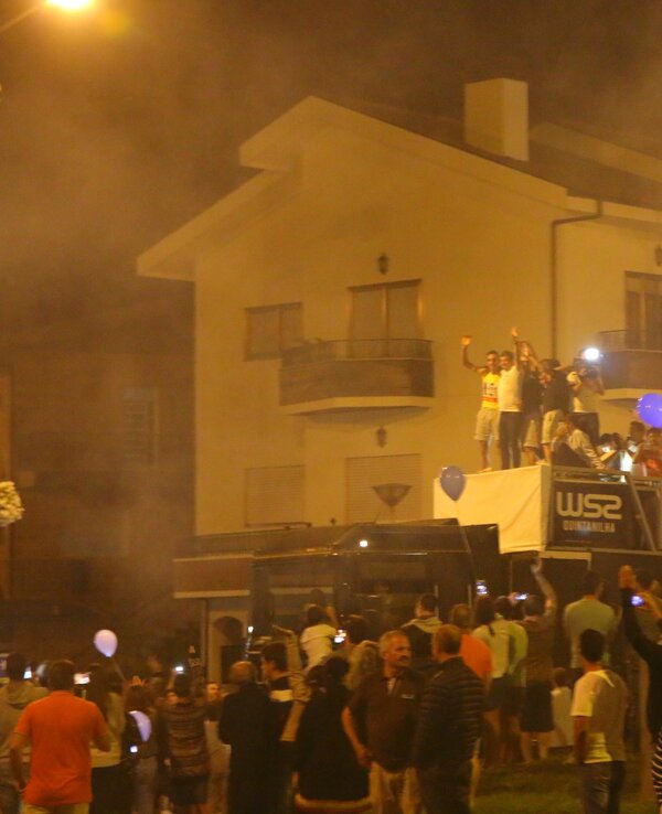Multidão festiva à noite, com balões e fumaça, celebrando em frente a um edifício.