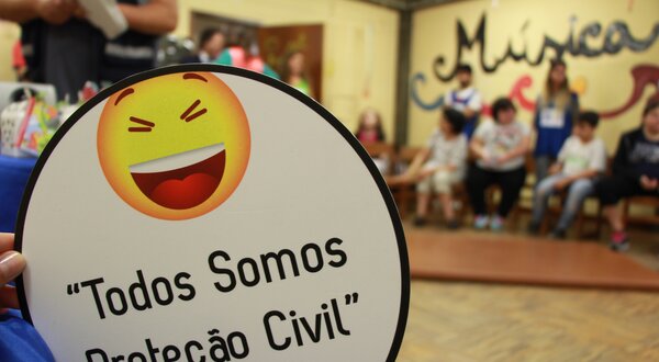 Placa com sorriso e frase "Todos Somos Proteção Civil" em evento.