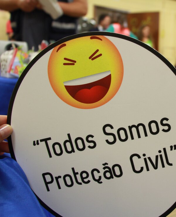 Placa com sorriso e frase "Todos Somos Proteção Civil" em evento.