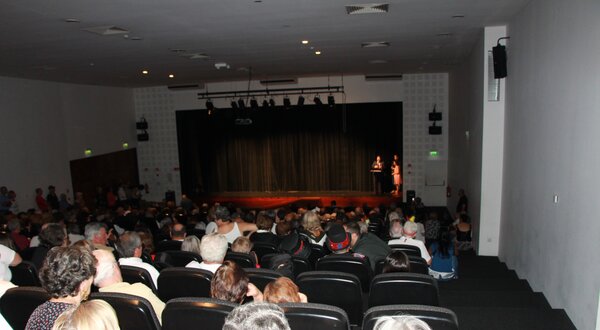 Público sentado em um auditório, assistindo a uma apresentação no palco.