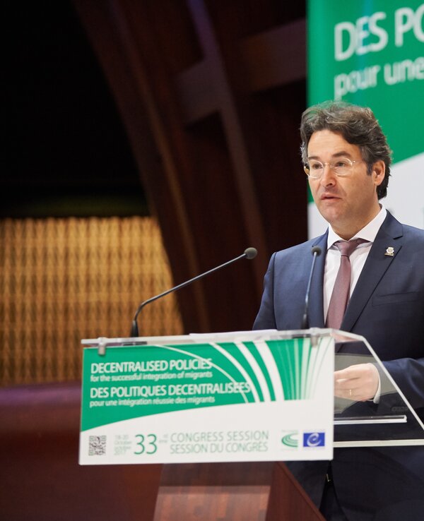 Orador em conferência, apresentando-se ao público com um painel atrás.