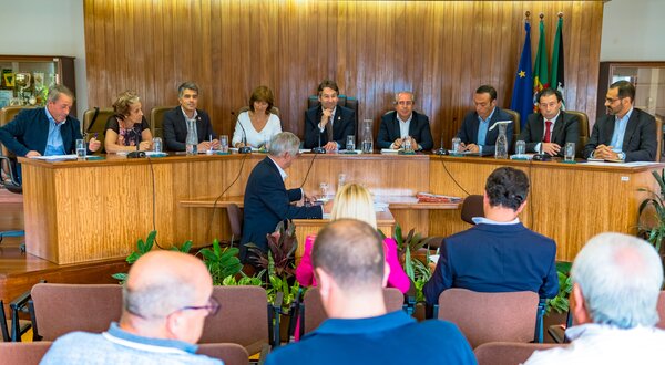 Reunião formal com várias pessoas sentadas à mesa, discutindo assuntos.