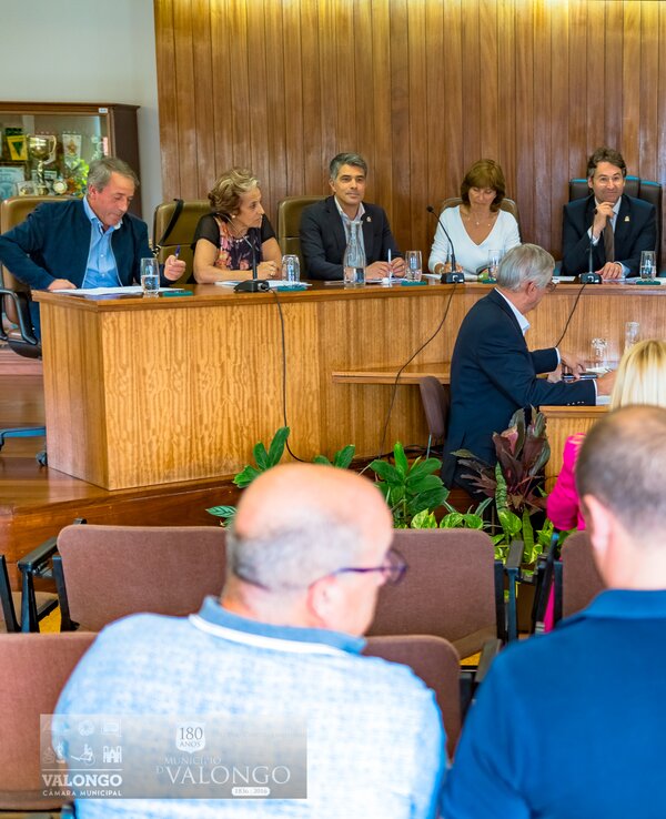 Reunião formal com várias pessoas sentadas à mesa, discutindo assuntos.