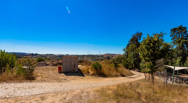 Caminho de terra com um contentor e vegetação sob um céu azul claro.