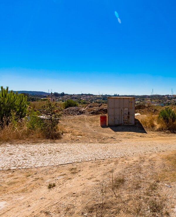 Caminho de terra com um contentor e vegetação sob um céu azul claro.