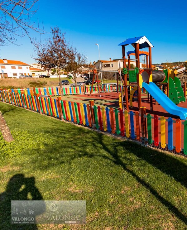 Parque infantil com escorrega e baloiços, cercado por uma vedação colorida.