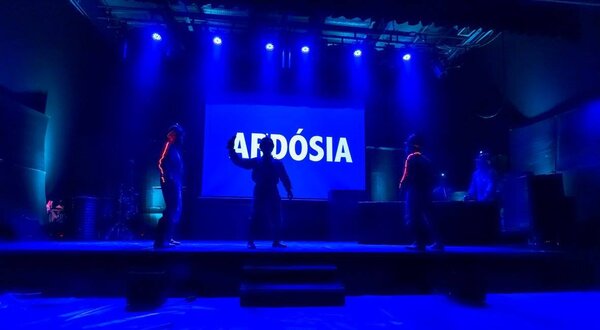 Três dançarinos em palco iluminado com luzes azuis, apresentando 'AFDÓSIA'.