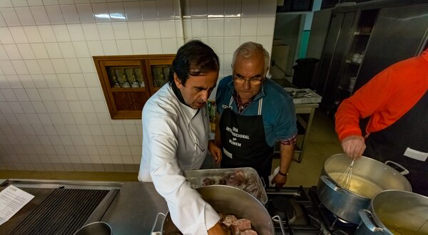 Dois homens a cozinhar em uma cozinha, um a adicionar temperos à carne.