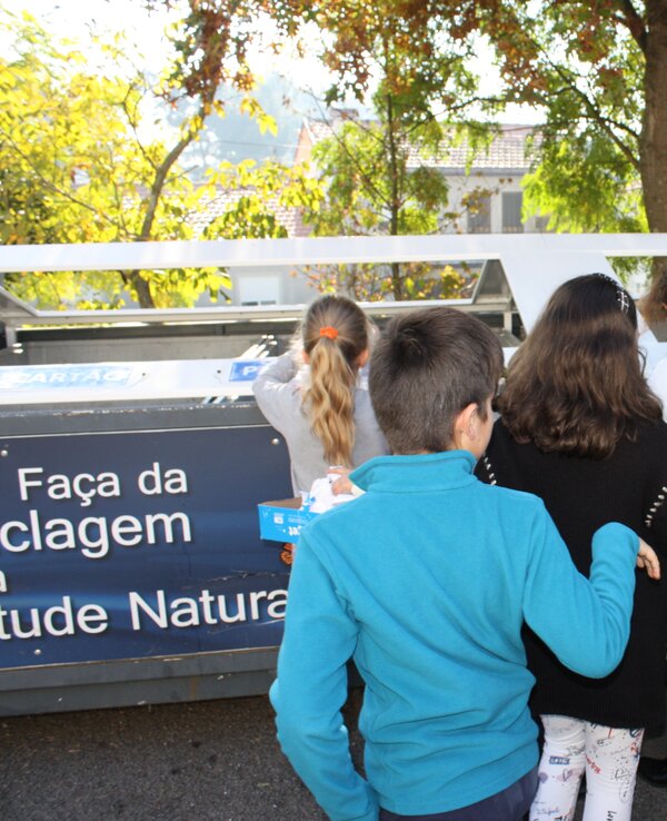 Grupo de crianças a reciclar em contentor, com árvores ao fundo.